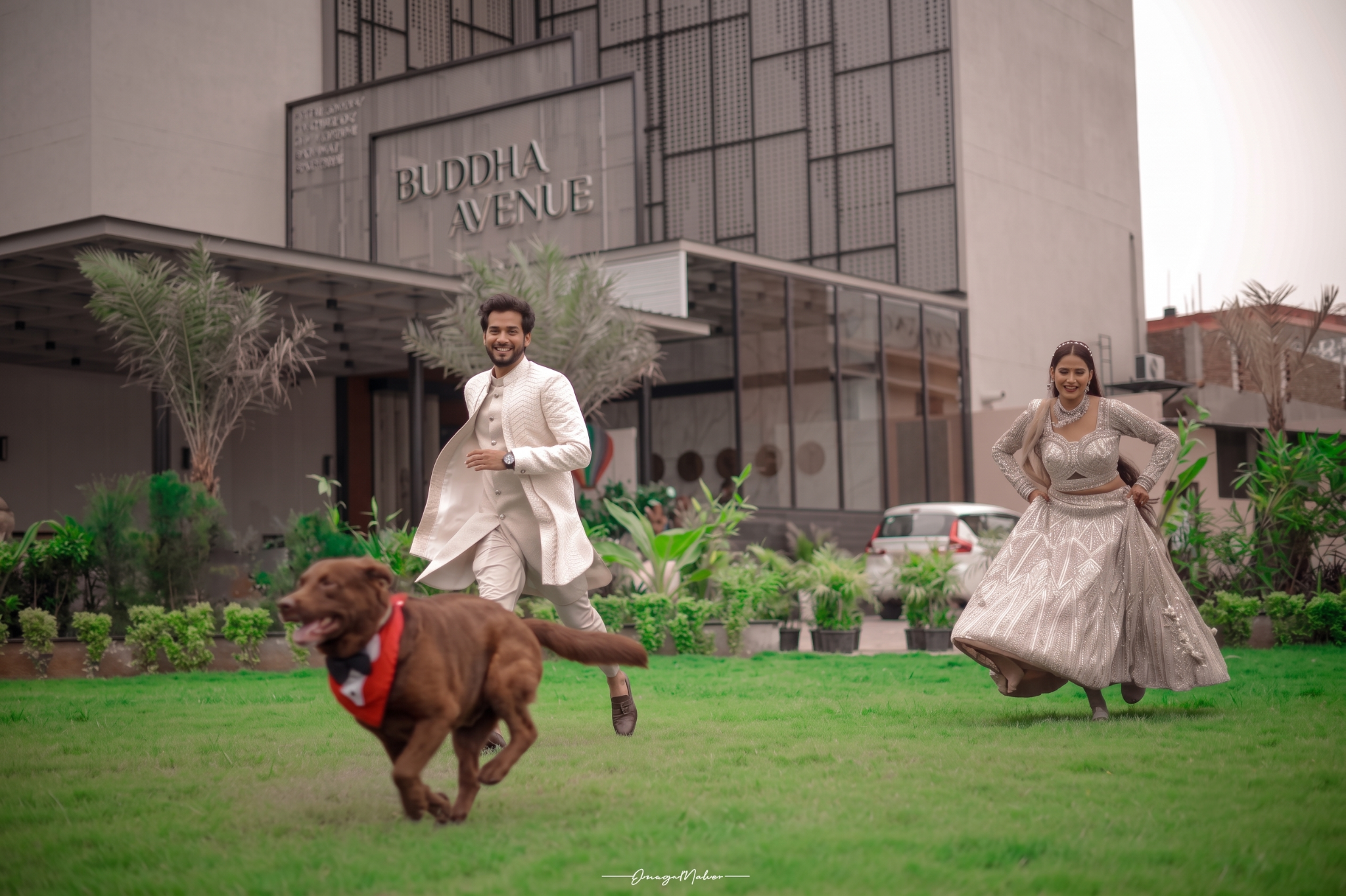 Pre-wedding couple running across a lawn with their dog outside a venue