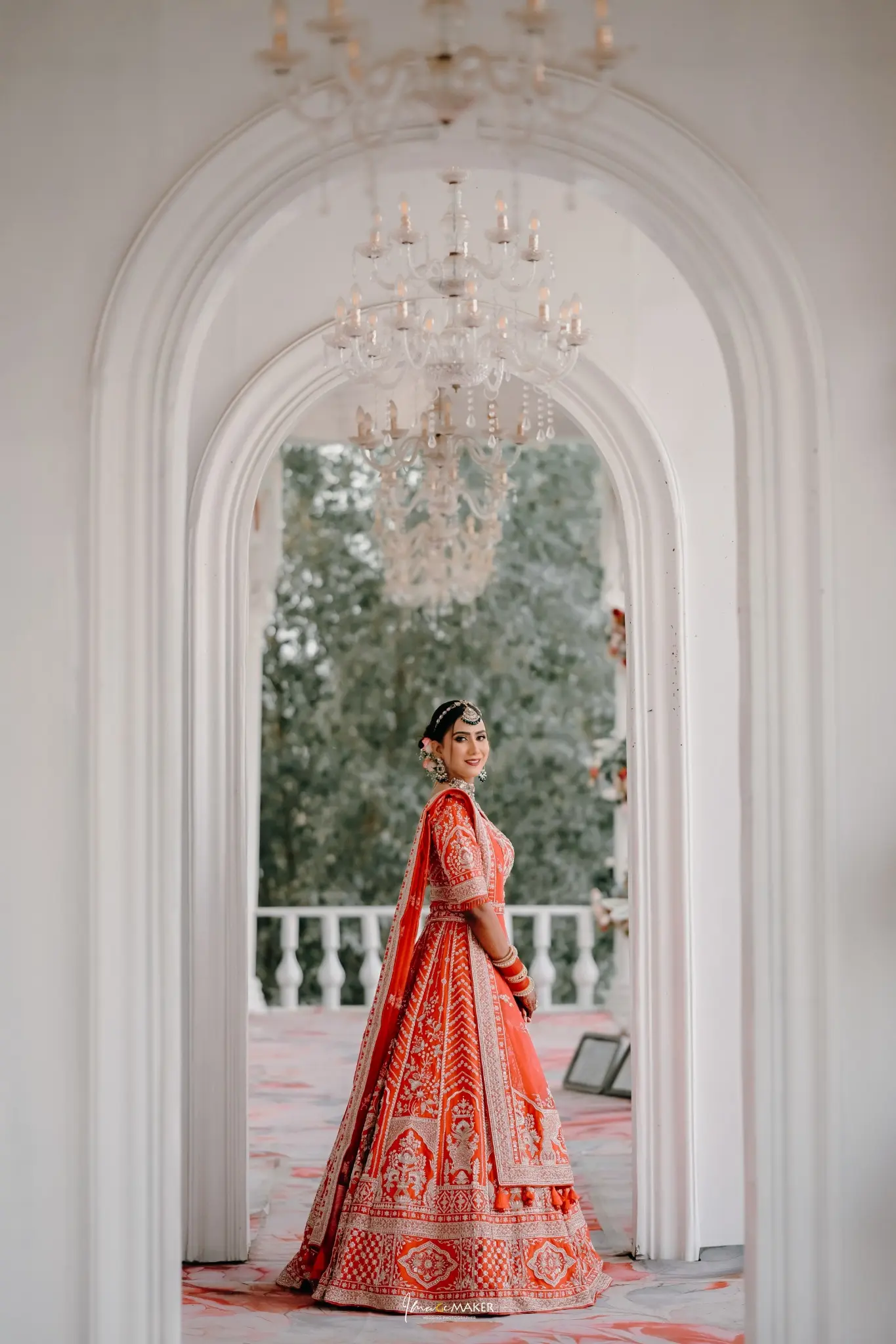 Bride in a red wedding lehenga standing beneath white arches and chandeliers